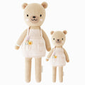 Goldie the honey bear in the regular and little sizes, shown from the front. Goldie is wearing a mushroom-shade apron dress and has a hand-knit flower bouquet stitched into her pocket.
