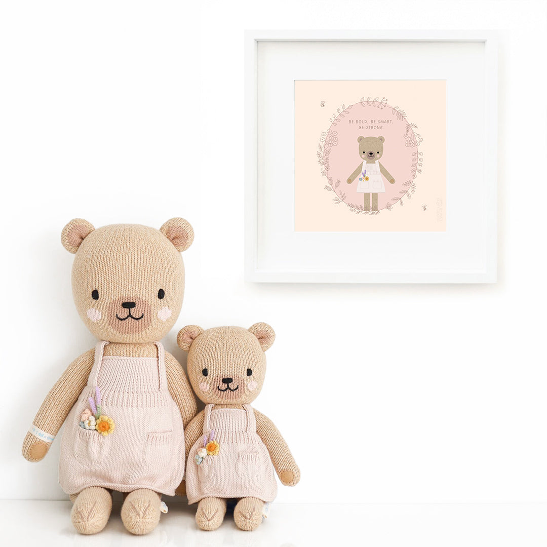 Two Goldie the honey bear stuffed dolls sitting beside a framed print with a picture of Goldie, with text that says "Be bold, be smart, be strong."