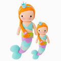 Isla the mermaid in the regular and little sizes, shown from the front. Isla has red hair that’s braided with a purple bow. She’s wearing a blue knitted crown, and the scales on her tail are green, purple and blue.