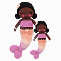 Maya the mermaid in the regular and little sizes, shown from the front. Maya has black hair with a pink shell on her head. She has a fringe, purple top, and the scales on her tail are purple, pink, peach and cream.