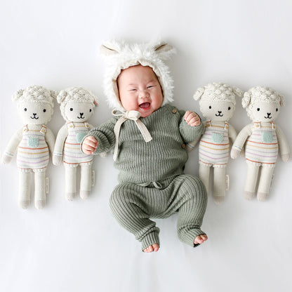 A laughing baby in a fluffy, lamb-eared bonnet, lying side-by-side with several Avery the lamb dolls.