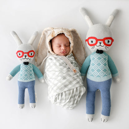 A sleeping baby in a bunny-eared bonnet lying side-by-side with two Benedict the bunny dolls in the regular and little sizes.