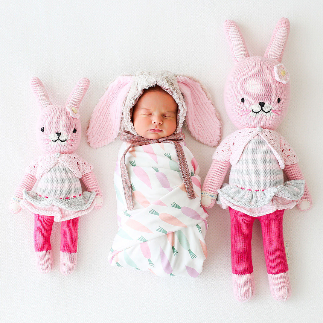 A baby in a bunny-eared bonnet lying between two Chloe the bunny dolls in the little and regular sizes.