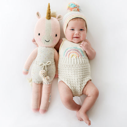 A smiling baby in a knit hat and romper with their arm around an Ella the unicorn stuffed doll.