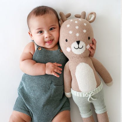A baby with their arm around an Elliott the fawn stuffed doll.
