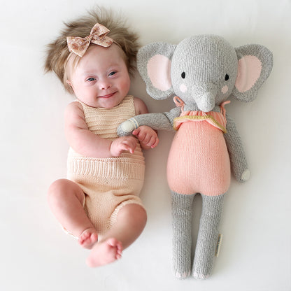 A smiling baby, wearing a sparkly hair bow, holding hands with Eloise the elephant. 