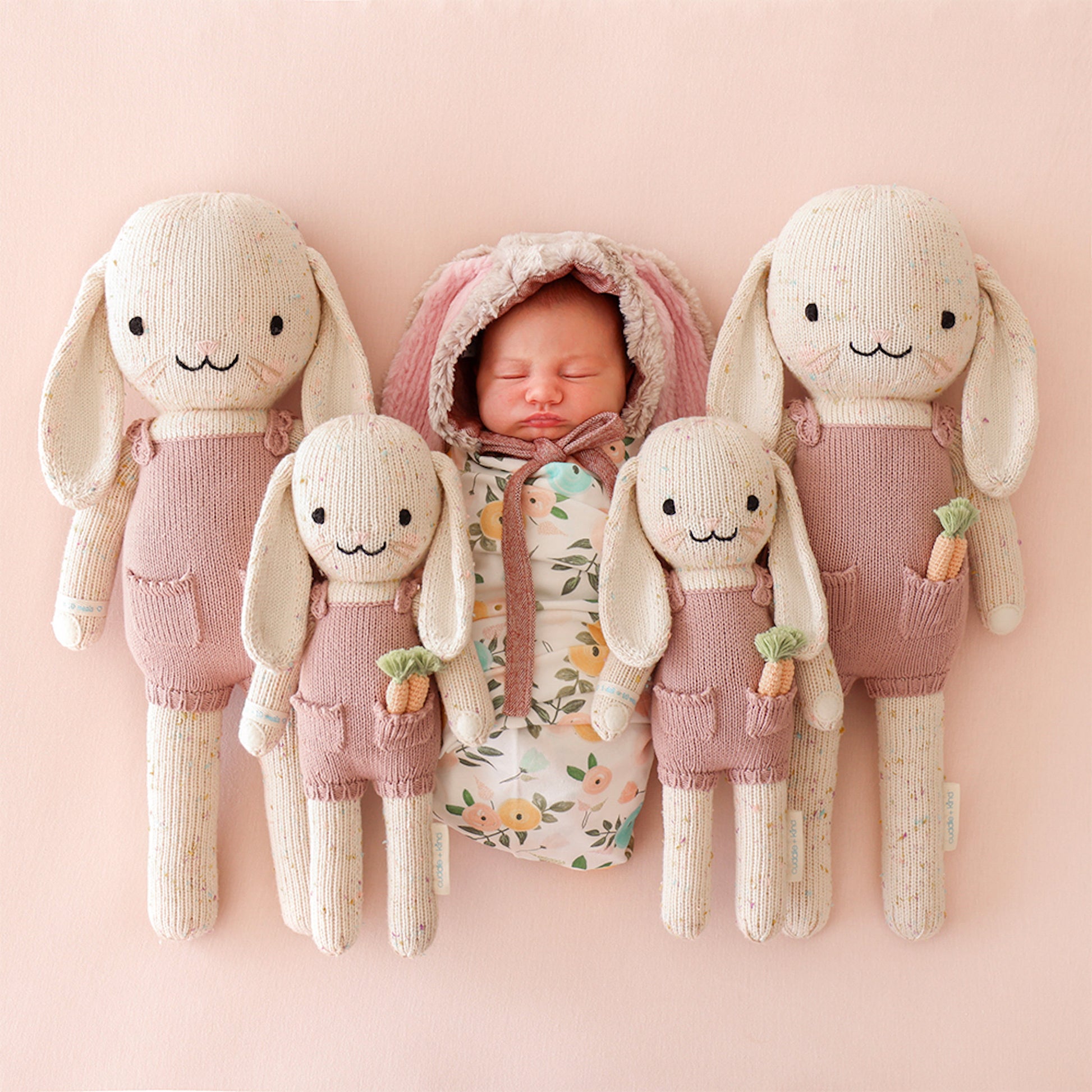 A baby, wearing a bunny-eared bonnet, snuggled between four Harper the bunny dolls in the little and regular sizes.