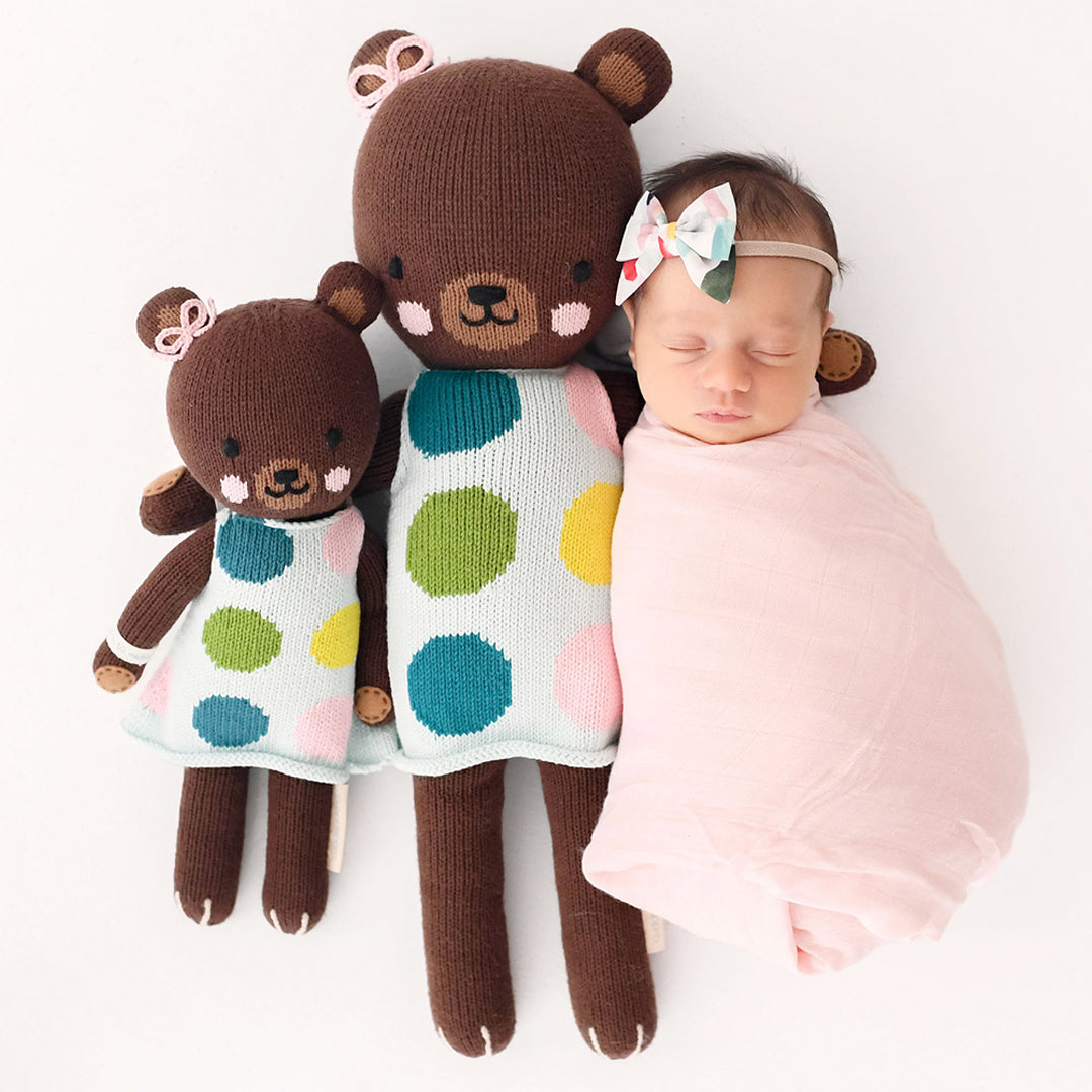 Two Ivy the bear, in the little and regular size, next to a sleeping, swaddled baby.