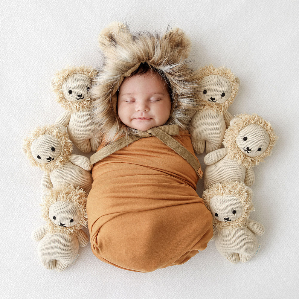 A sleeping baby in a fluffy lion hood snuggled with six baby lion stuffed animals.