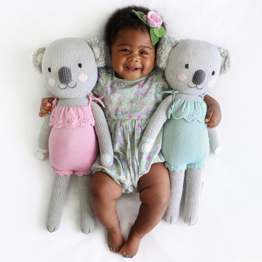 A smiling baby with two Claire the koala stuffed dolls in orchid and mint.