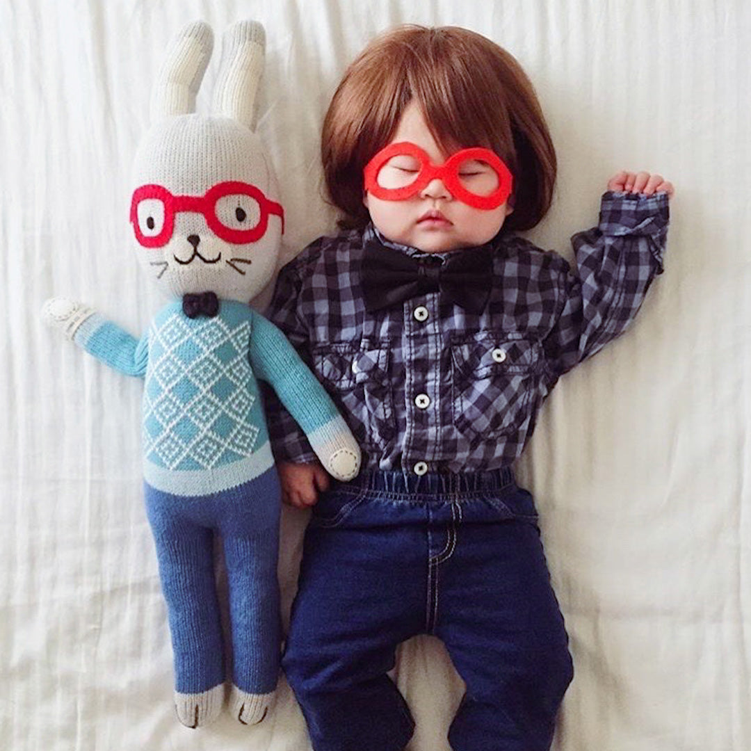 A sleeping baby lying next to Benedict the bunny. The baby is wearing red felt glasses and a black bow-tie to match Benedict.
