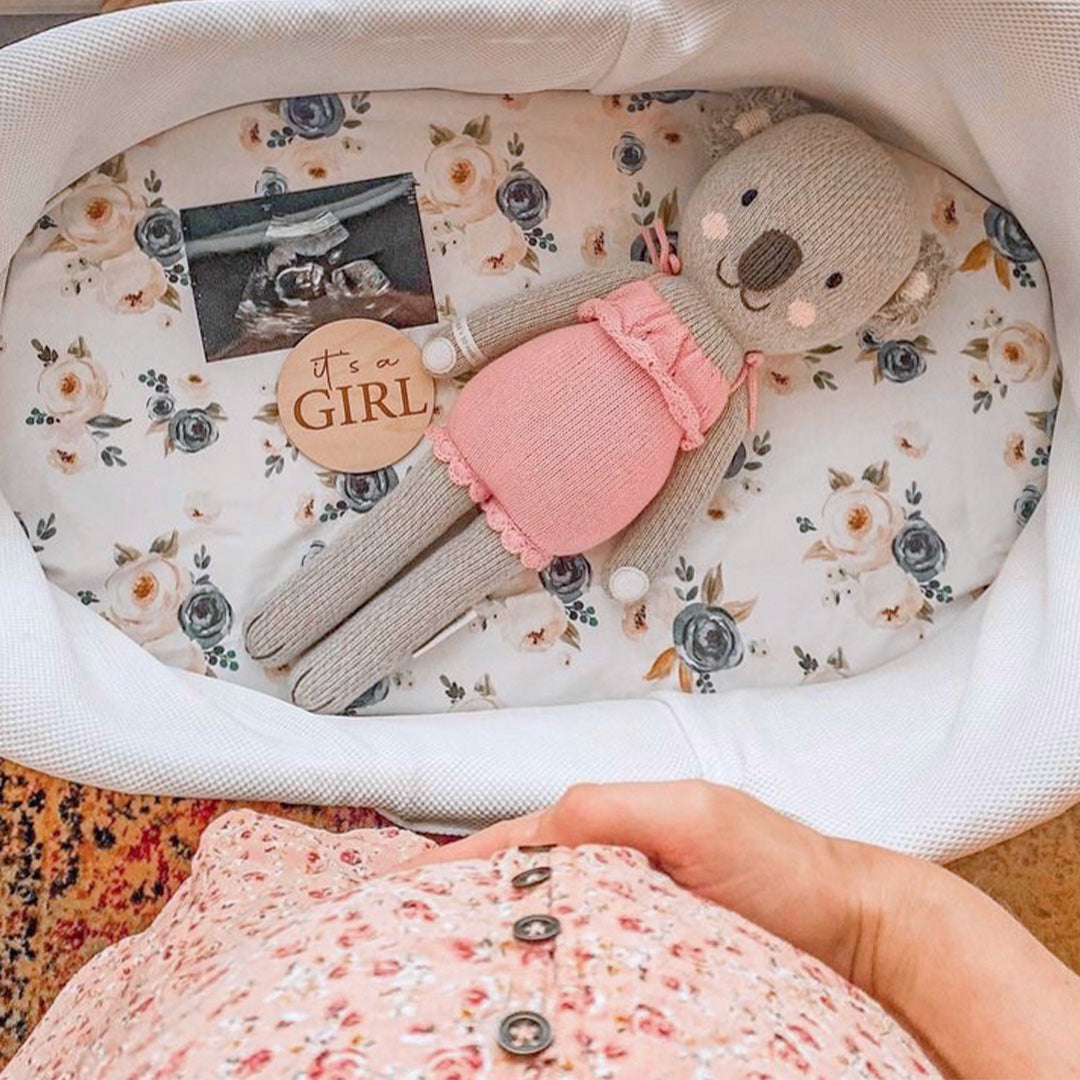 Claire the koala inside a bassinet alongside an ultrasound image and a round wooden sign that says “it's a girl!" A woman holding her baby bump can be seen beside the bassinet. 