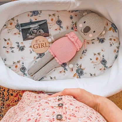 Claire the koala inside a bassinet alongside an ultrasound image and a round wooden sign that says “it's a girl!" A woman holding her baby bump can be seen beside the bassinet. 