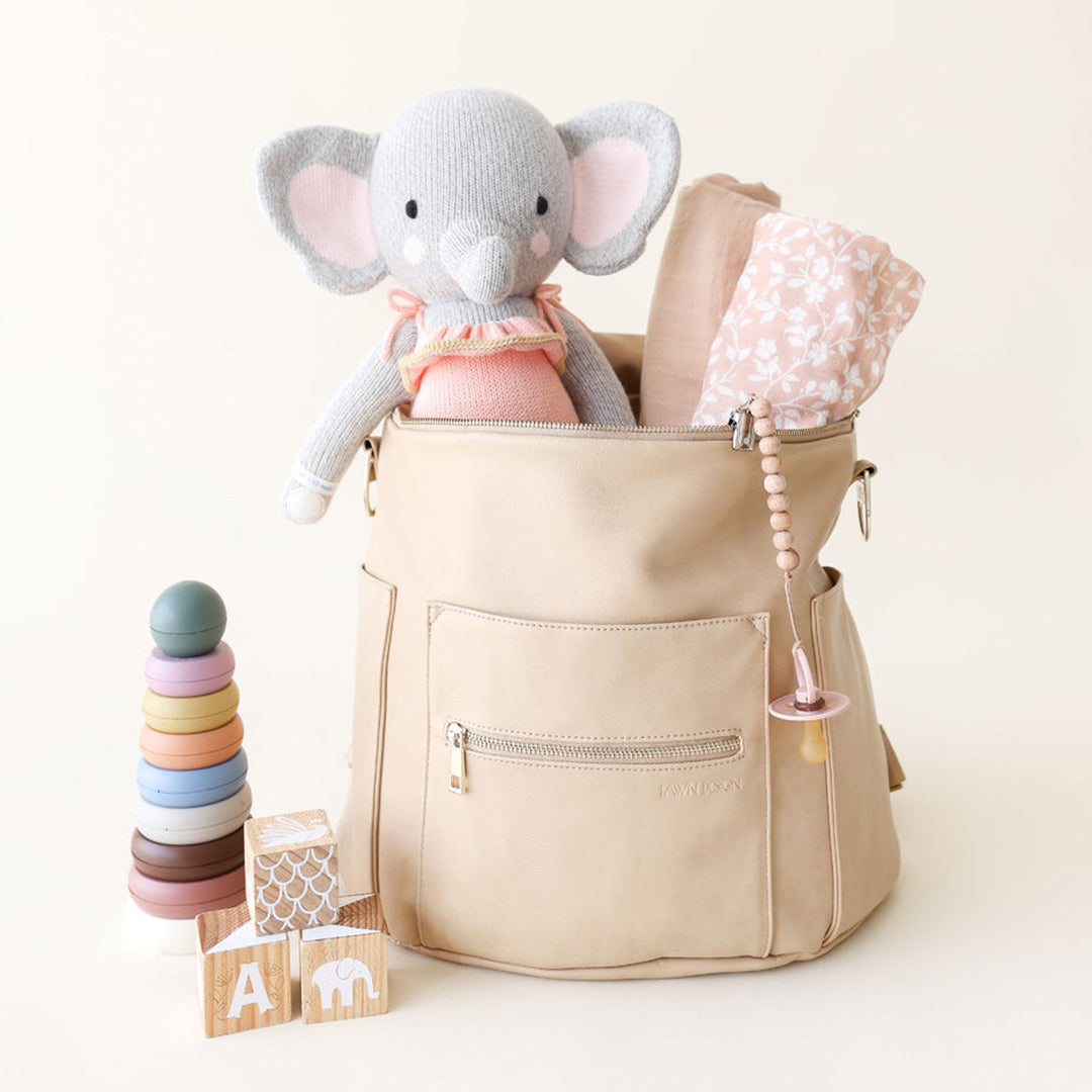 Eloise the elephant sitting in a baby bag, alongside two rolled receiving blankets and a soother. Beside the bag are children's toys.