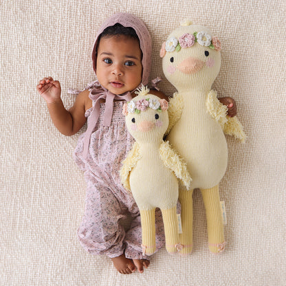 A baby, wearing a knit bonnet, with their arm around two Flora the duckling stuffed dolls, in the regular and little size.