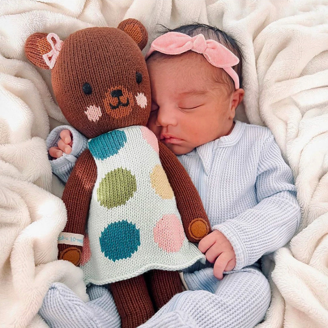 A sleeping baby, wearing a pink bow, snuggling an Ivy the bear stuffed doll.