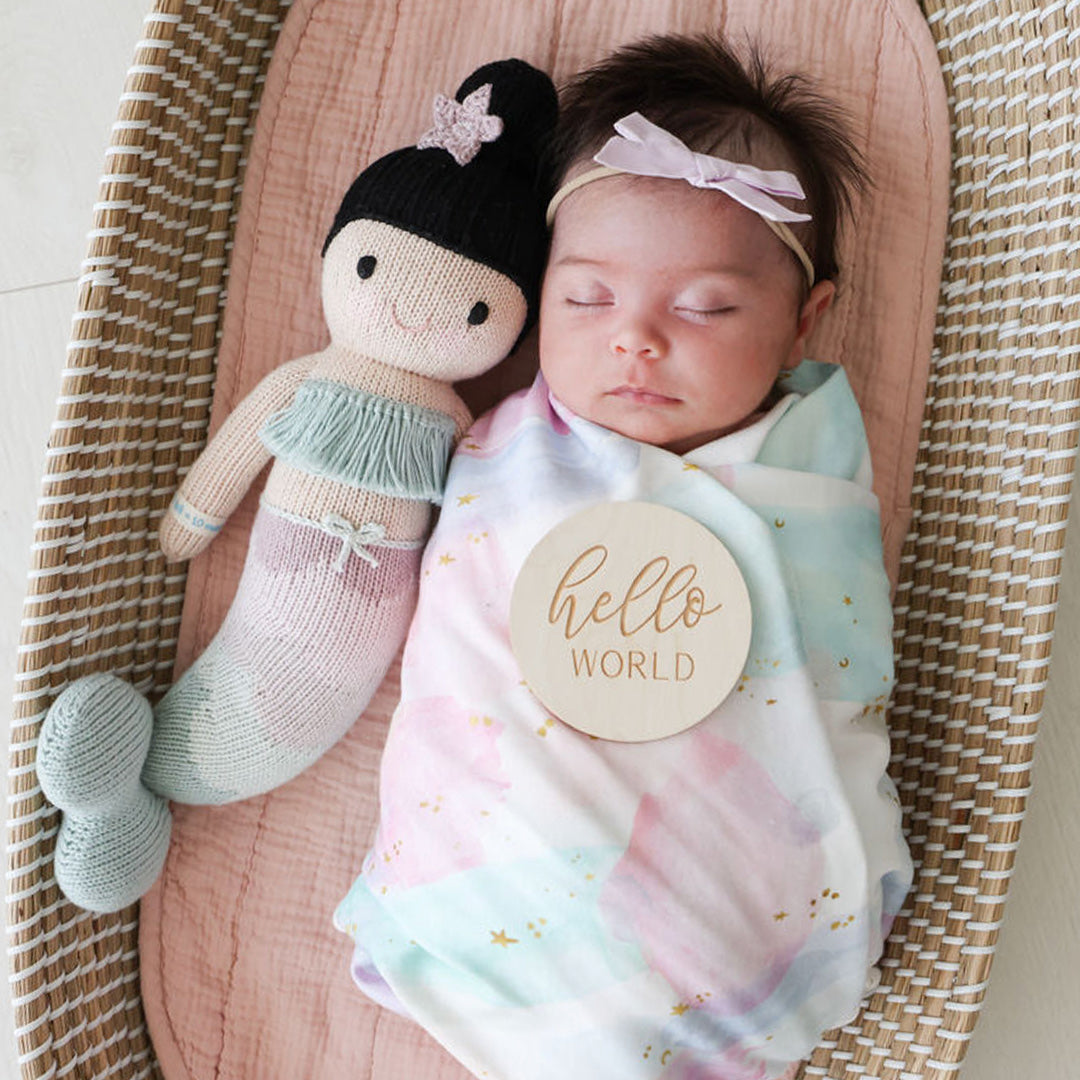 A sleeping baby in a bassinet, snuggled beside Luna the mermaid. There is a small, wooden sign resting on the baby with text that says, “hello world.”