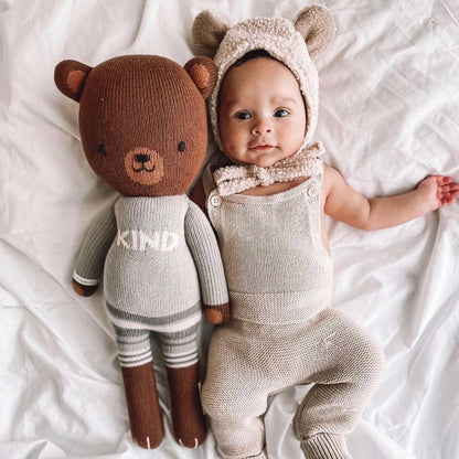 A baby in a bear-eared bonnet with their arm around Oliver the bear.