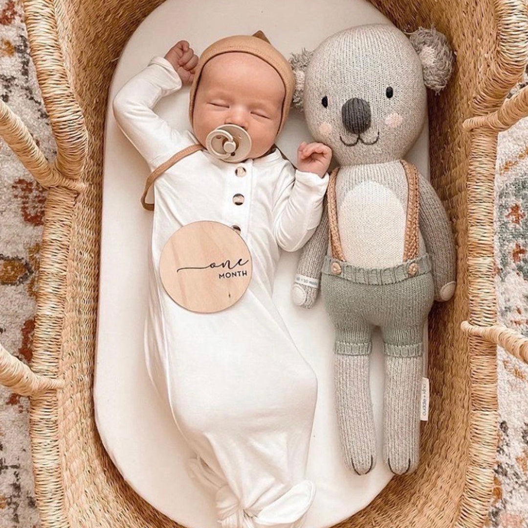A sleeping baby, stretched out in a bassinet beside Quinn the koala. A little wooden sign resting on the baby’s tummy has text that says “one month.”