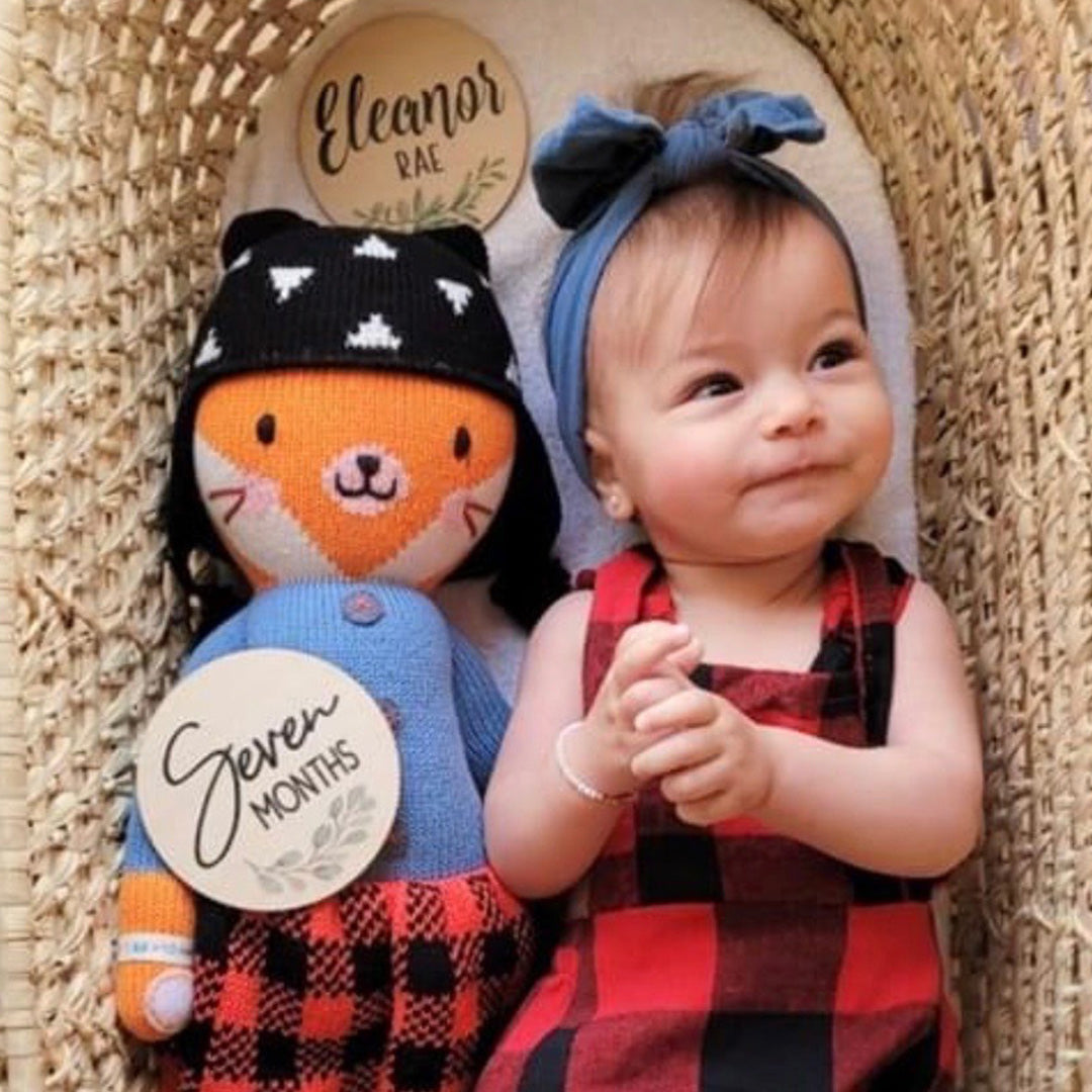 A smiling baby, wearing a plaid romper, with a Sadie the fox stuffed doll.