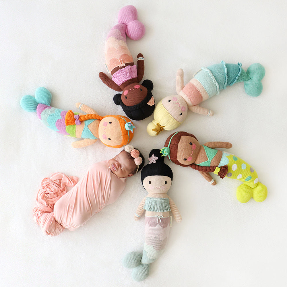 A sleeping baby lying in a circle head-to-head with five mermaid dolls: Isla, Maya, Skye, Pearl and Luna.