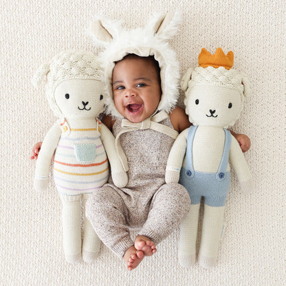 A baby in a fluffy lamb-eared bonnet lying side by side with two lamb dolls from cuddle and kind: Avery and Sebastian.