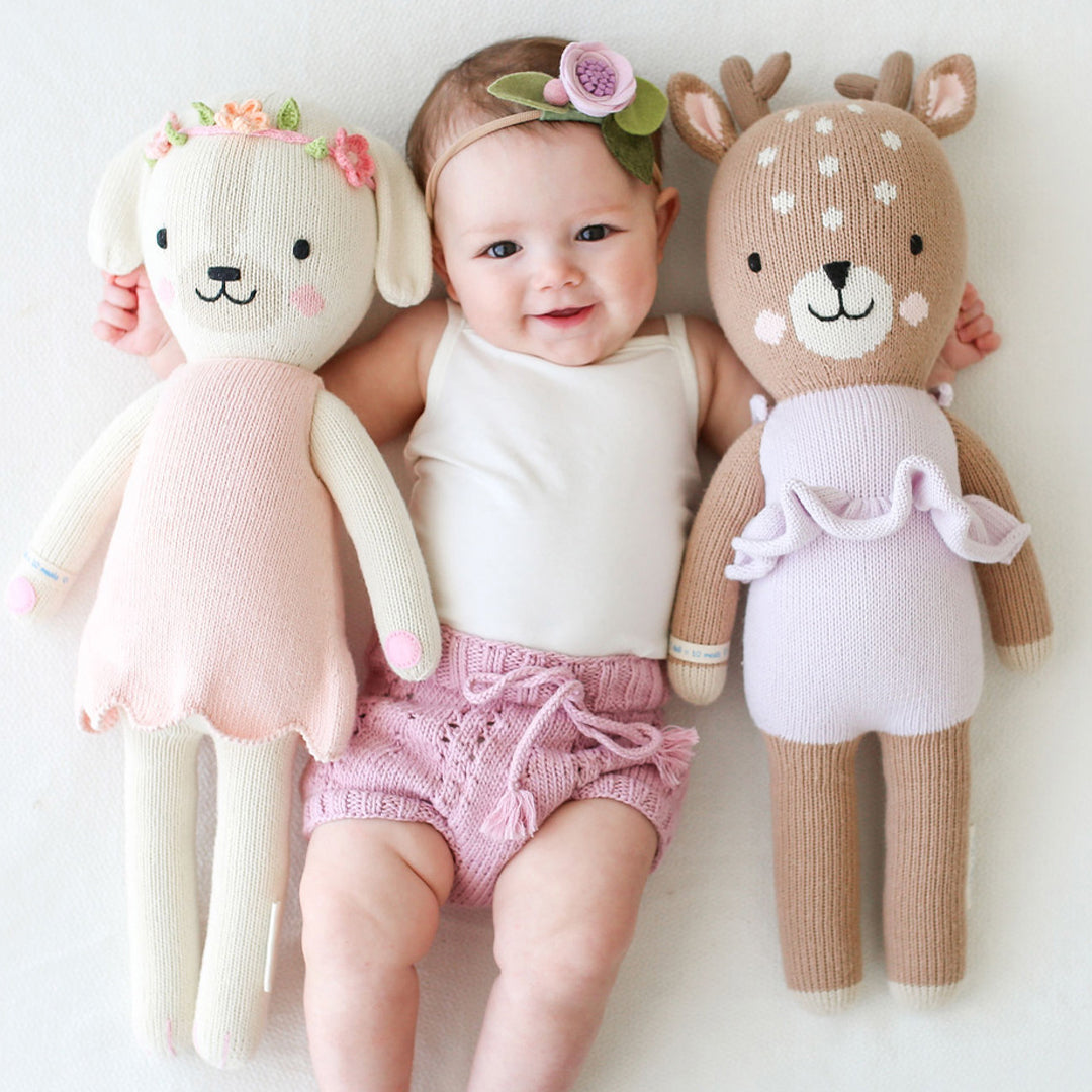 A smiling baby wearing a floral headband with their arms around a Charlotte the dog stuffed doll and a Violet the fawn stuffed doll. 