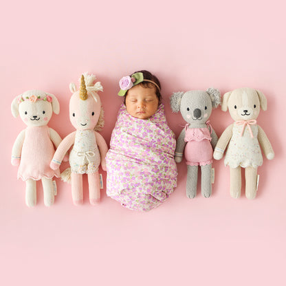 A sleeping baby wrapped snugly in a receiving blanket, surrounded by four cuddle and kind hand-knit stuffed dolls.