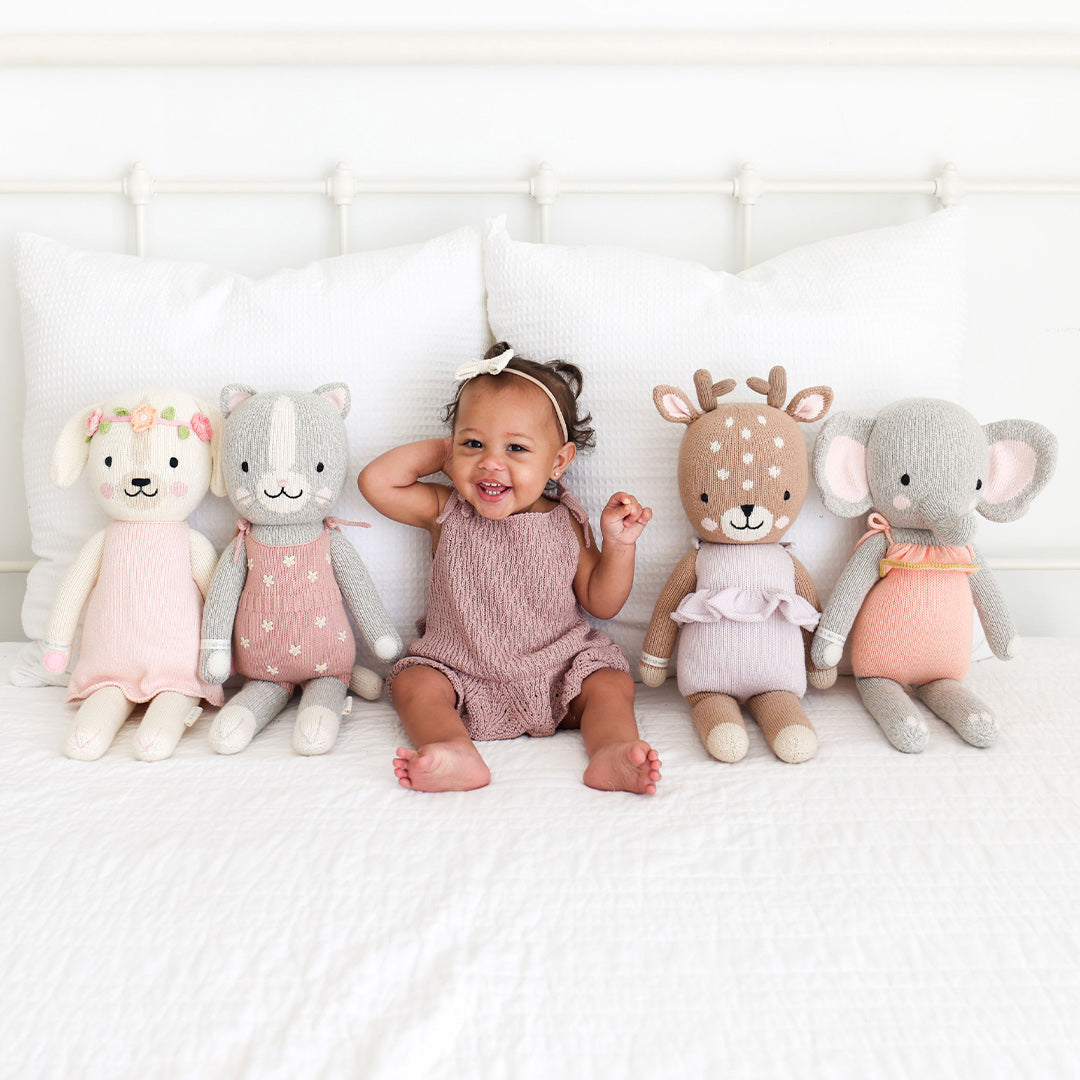 A laughing toddler sitting in the center of four cuddle and kind hand-knit stuffed dolls.