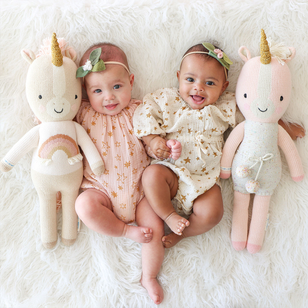 Two smiling babies with their arms around one Ella the unicorn and one Zara the unicorn stuffed doll.