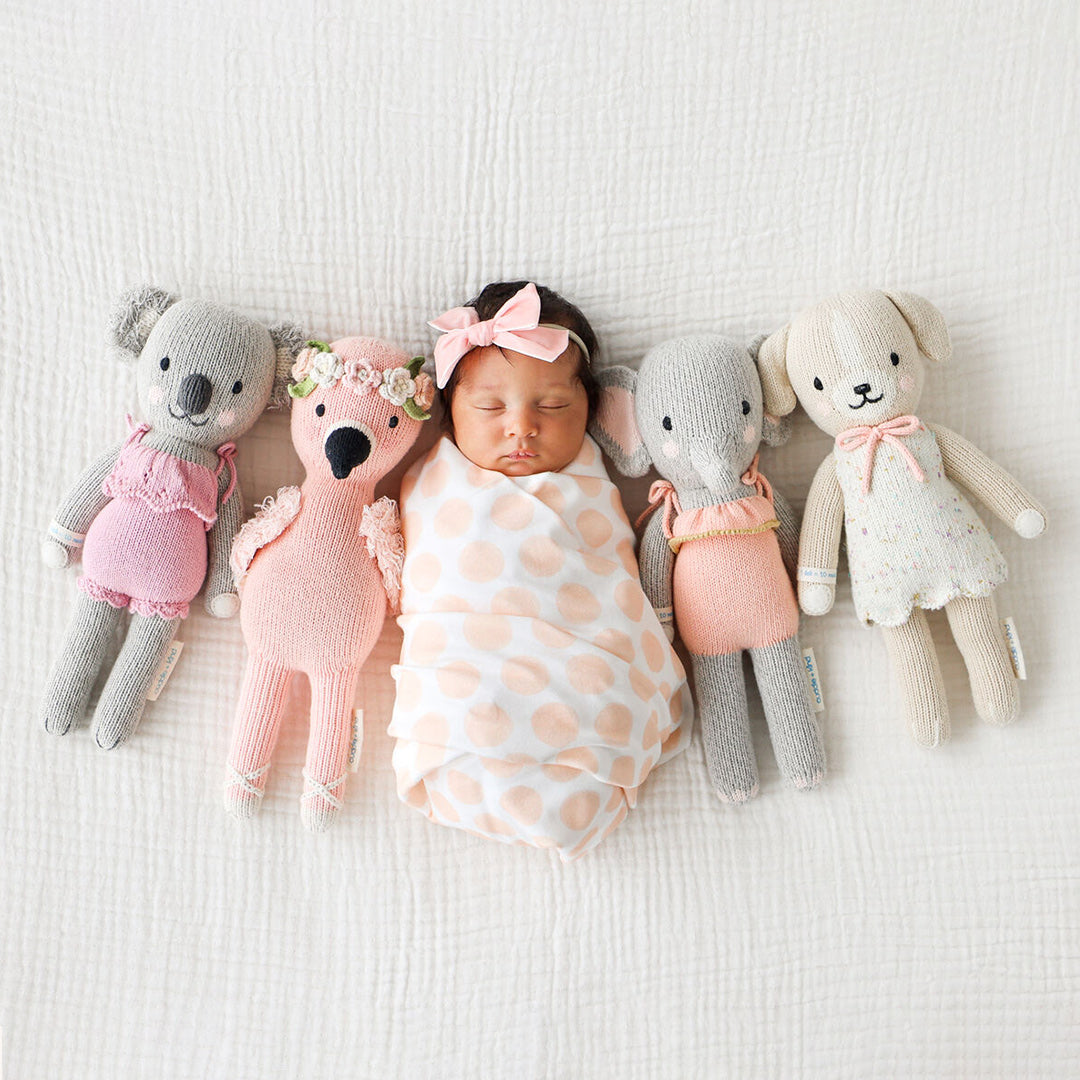 A sleeping baby, in a pink hair bow, surrounded by four cuddle and kind stuffed dolls: Claire the koala, Penelope the flamingo, Eloise the elephant and Mia the dog.