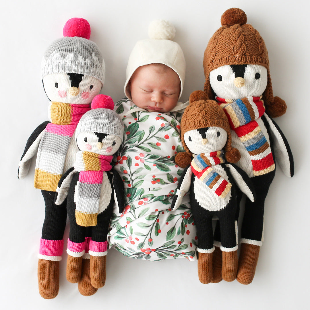A sleeping baby lying in between two Everest the penguin stuffed dolls and two Aspen the penguin stuffed dolls in the regular and little sizes.