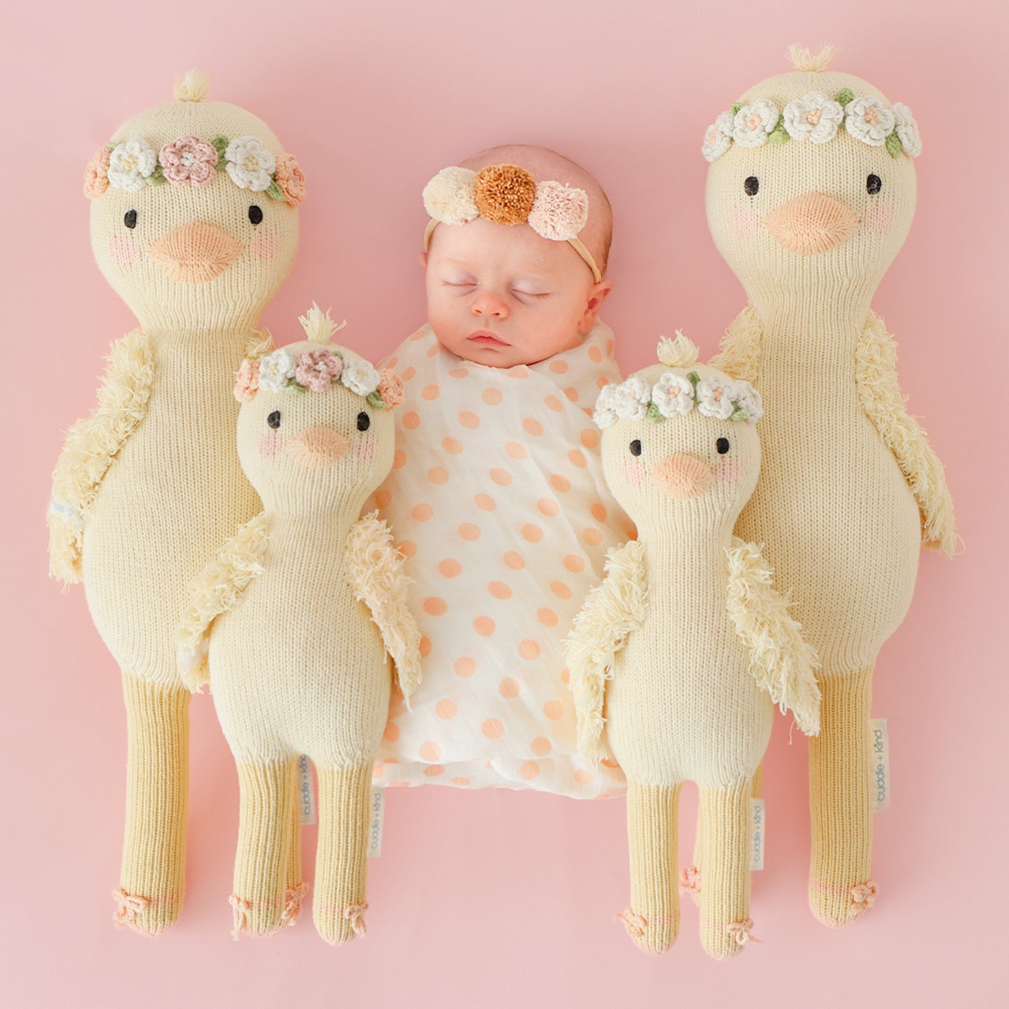 A sleeping baby, wearing a pom pom headband, nestled between four Flora the duckling stuffed dolls, in the regular and little sizes.