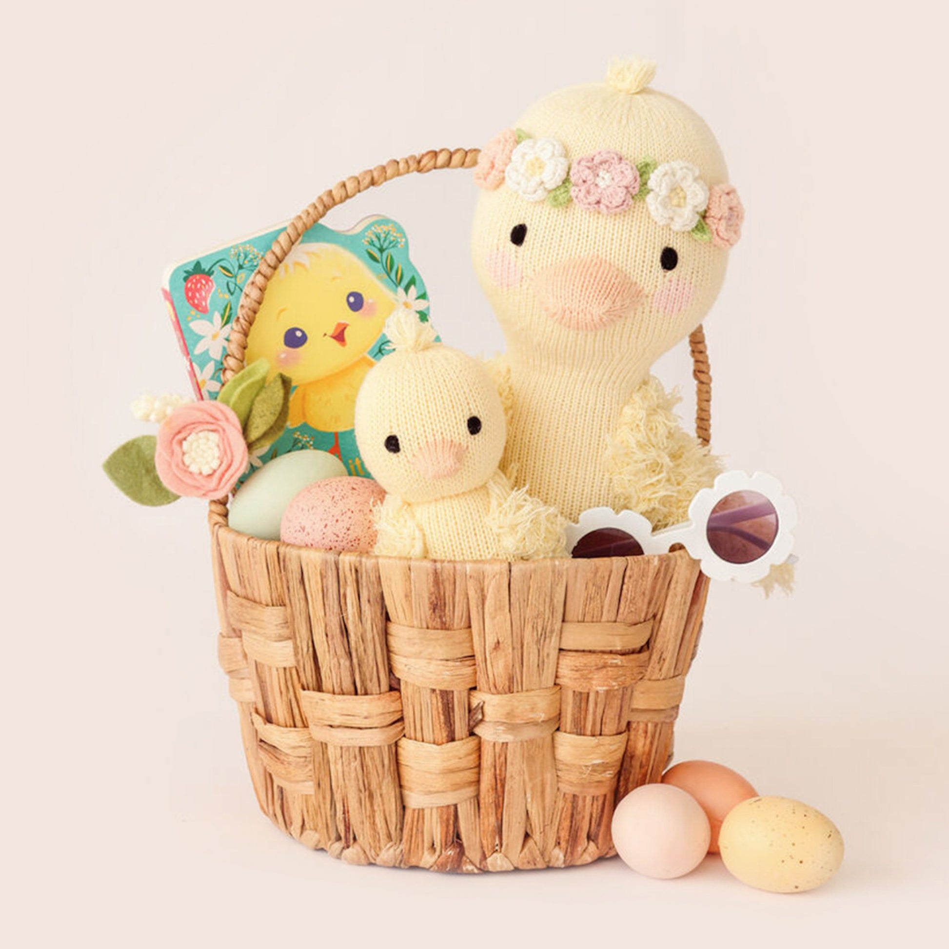 A Flora the duckling stuffed doll and a baby duckling stuffed animal in a basket, along with pastel eggs, a children's book and a pair of flower-shaped sunglasses.