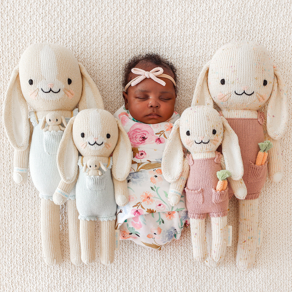 A sleeping baby, wrapped in a swaddle, surrounded by four cuddle+kind stuffed dolls.