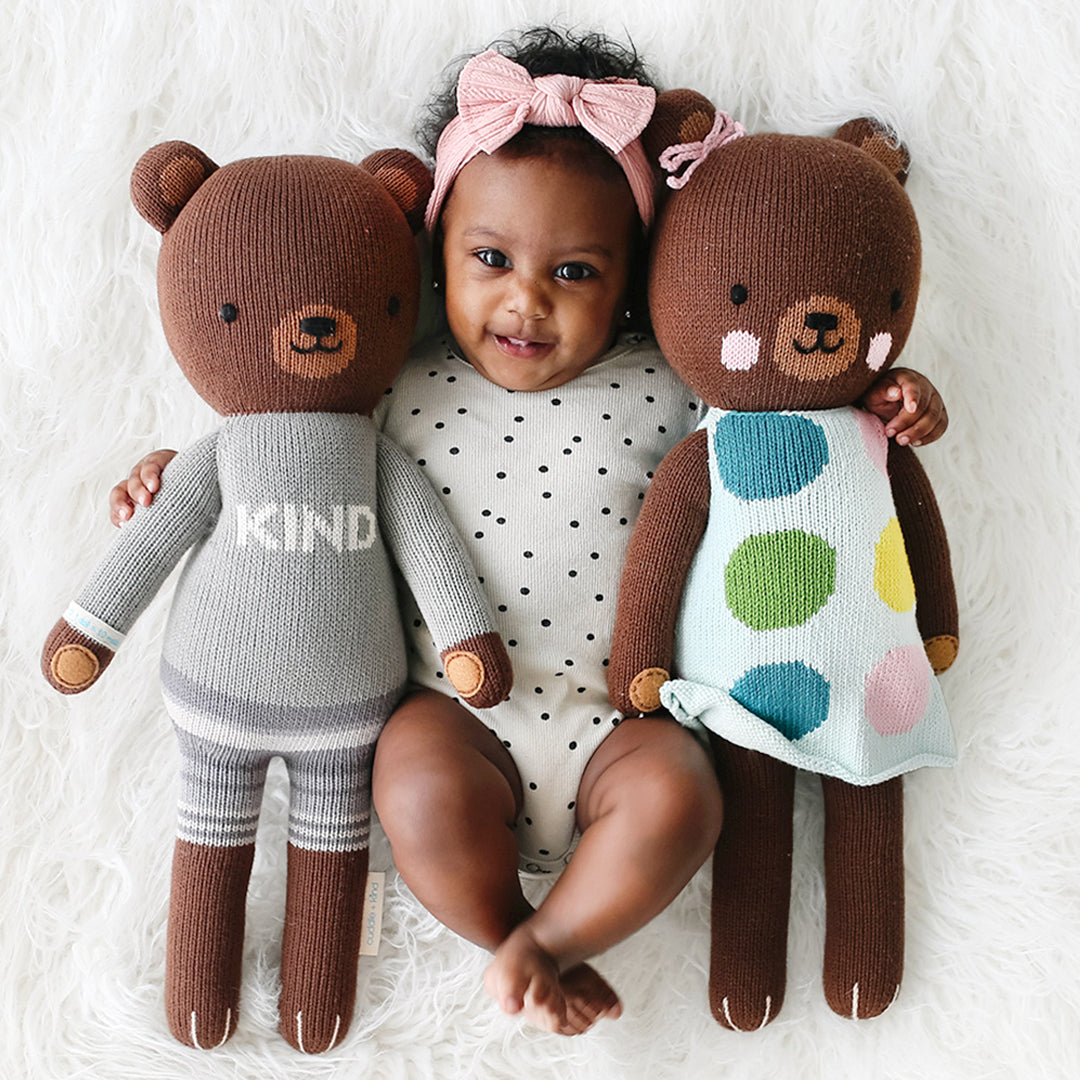 A smiling baby with their arms around two cuddle+kind stuffed dolls, Oliver and Ivy the bears.