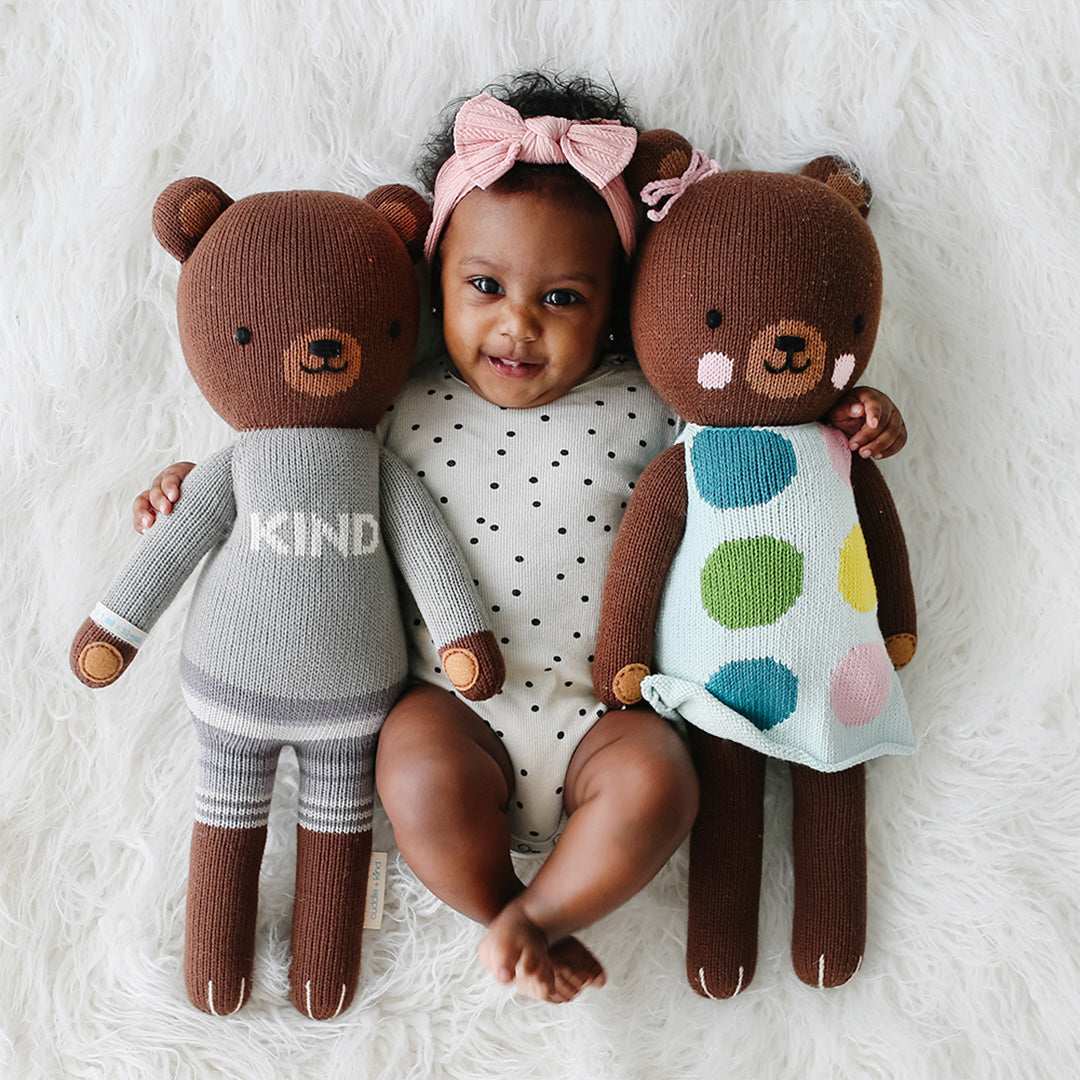 A smiling baby with her arms around cuddle+kind stuffed bears, Oliver and Ivy.
