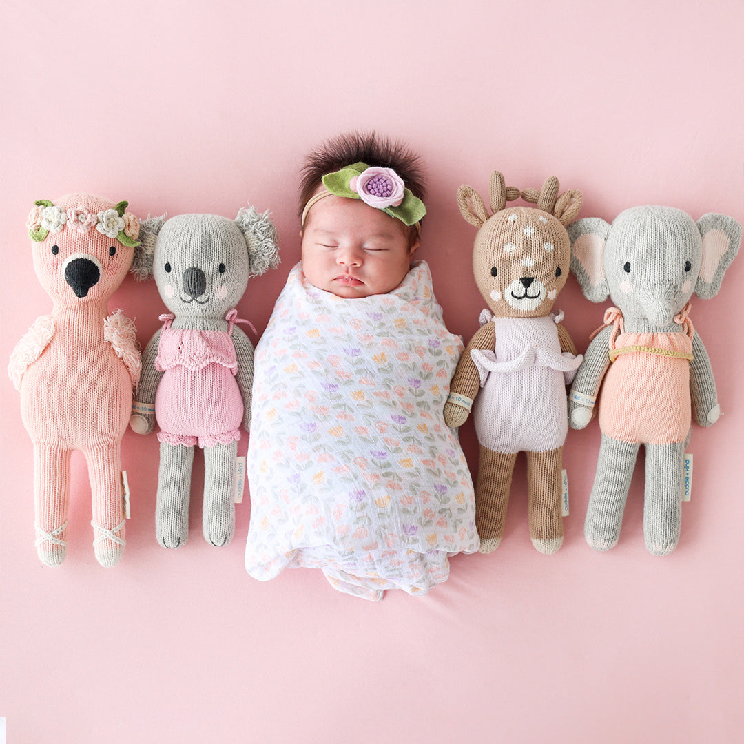 A sleeping, swaddled baby, lying in the middle of four cuddle+kind stuffed dolls: Penelope, Claire, Violet and Eloise.