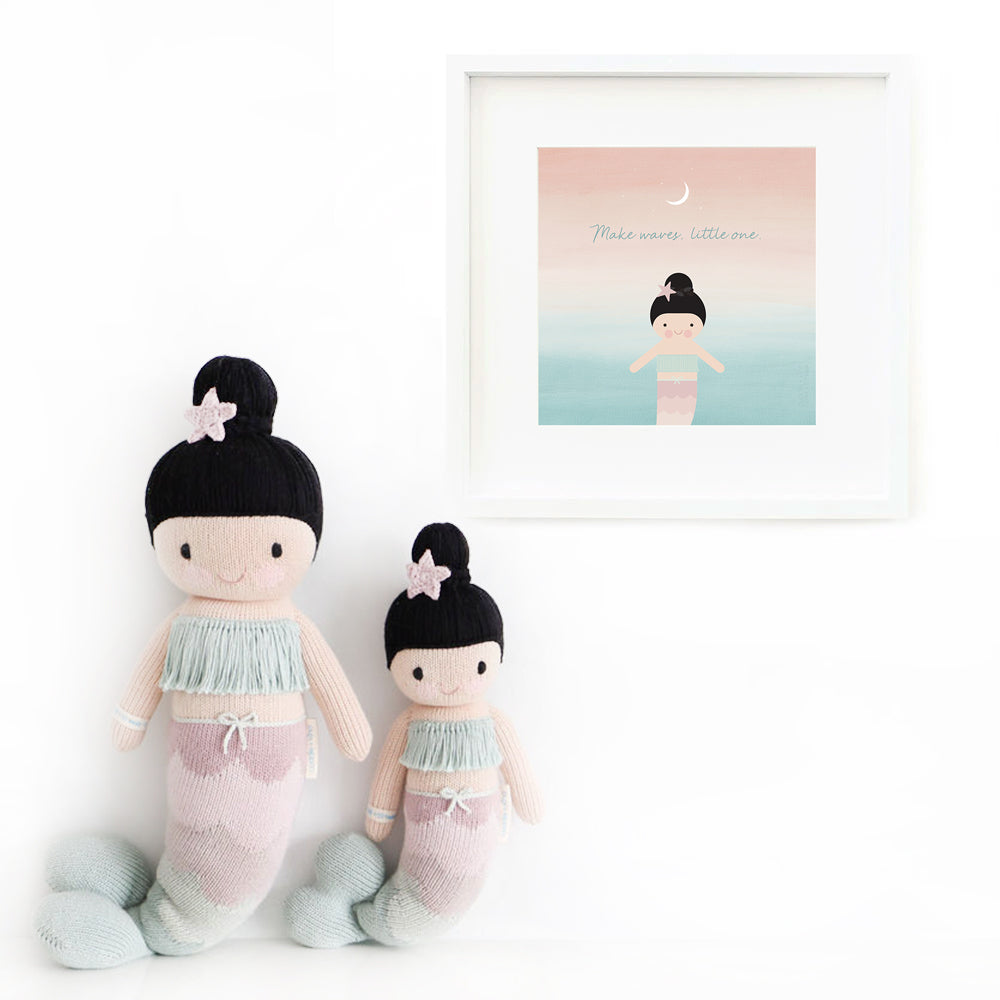 Two Luna the mermaid stuffed dolls sitting beside a framed print with a picture of Luna that says “Make waves, little one.”