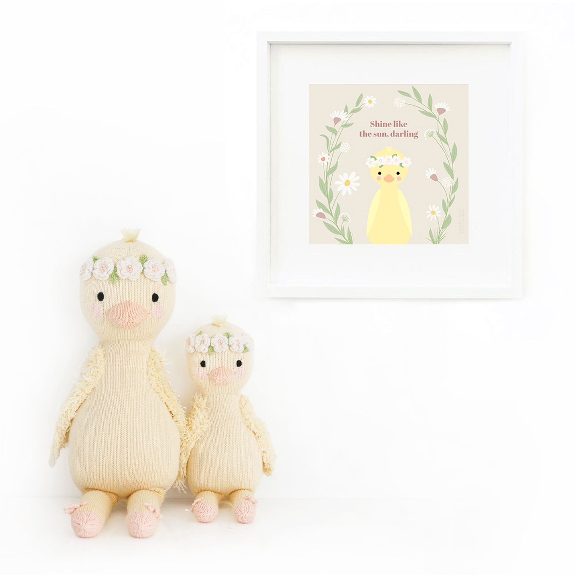Two Flora the duckling stuffed dolls sitting beside a framed print with a drawing of Flora, with text that says “Shine like the sun, darling.”