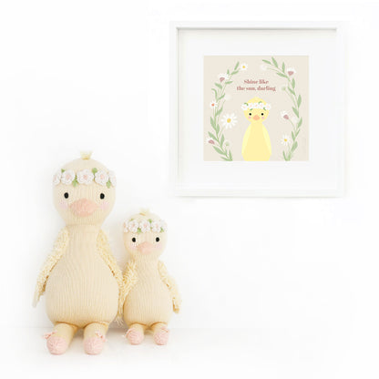 Two Flora the duckling stuffed dolls sitting beside a framed print with a drawing of Flora, with text that says “Shine like the sun, darling.”