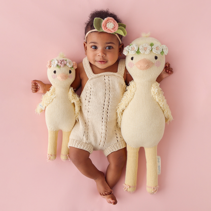 A smiling baby, wearing a floral headband, with their arms around two Flora the duckling stuffed dolls, in the regular and little size.