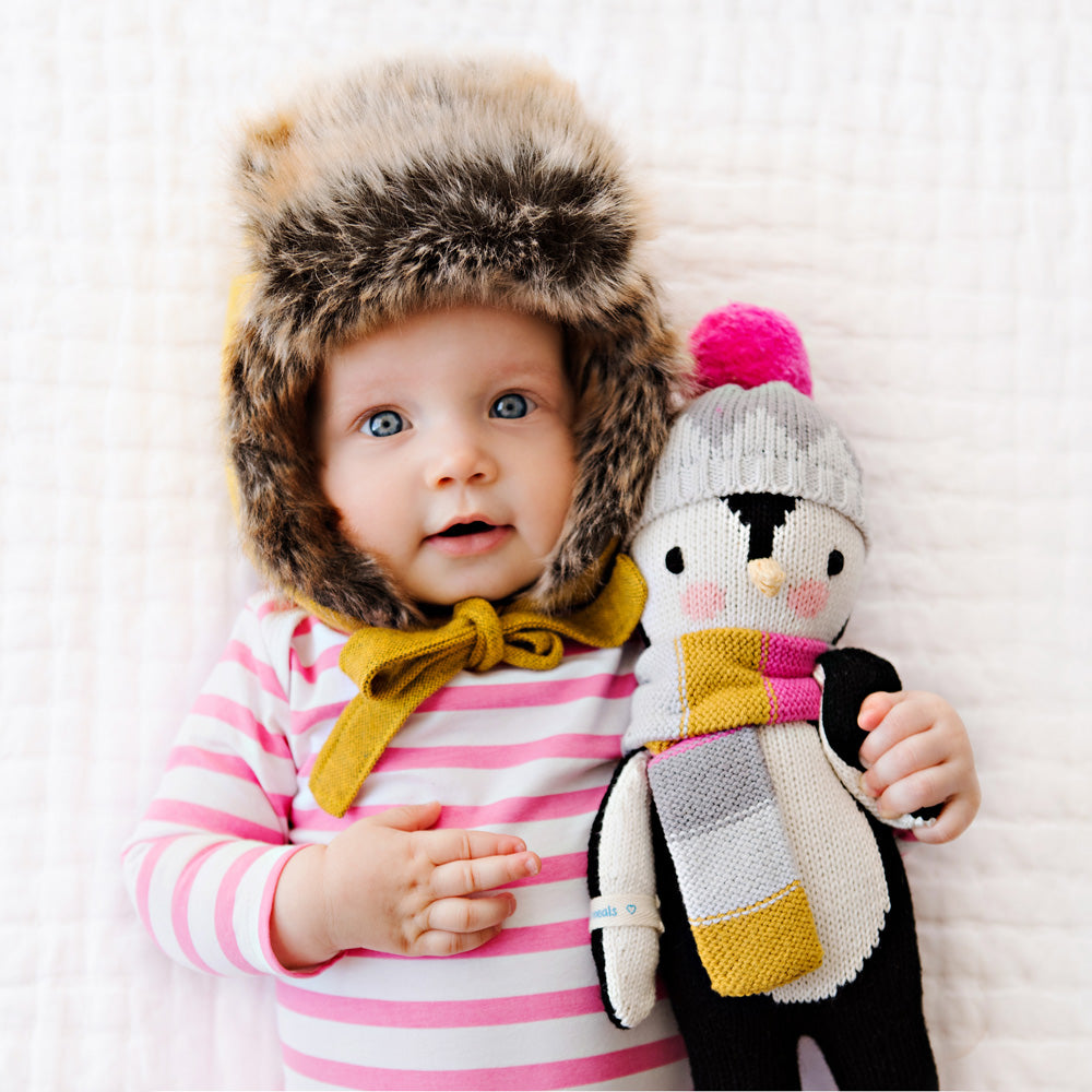 A baby in a warm winter hat hugging their Aspen the penguin stuffed doll.