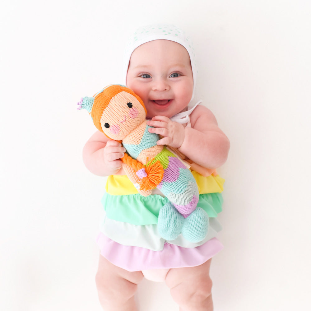 A laughing baby holding an Isla the mermaid doll. The baby’s outfit has rainbow ruffles to match Isla’s tail.
