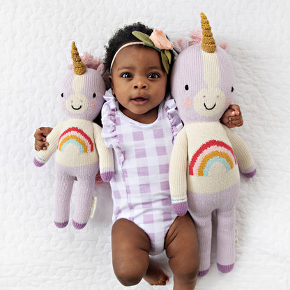A baby with her arms around two Zoe the unicorn dolls, in the little and regular sizes.