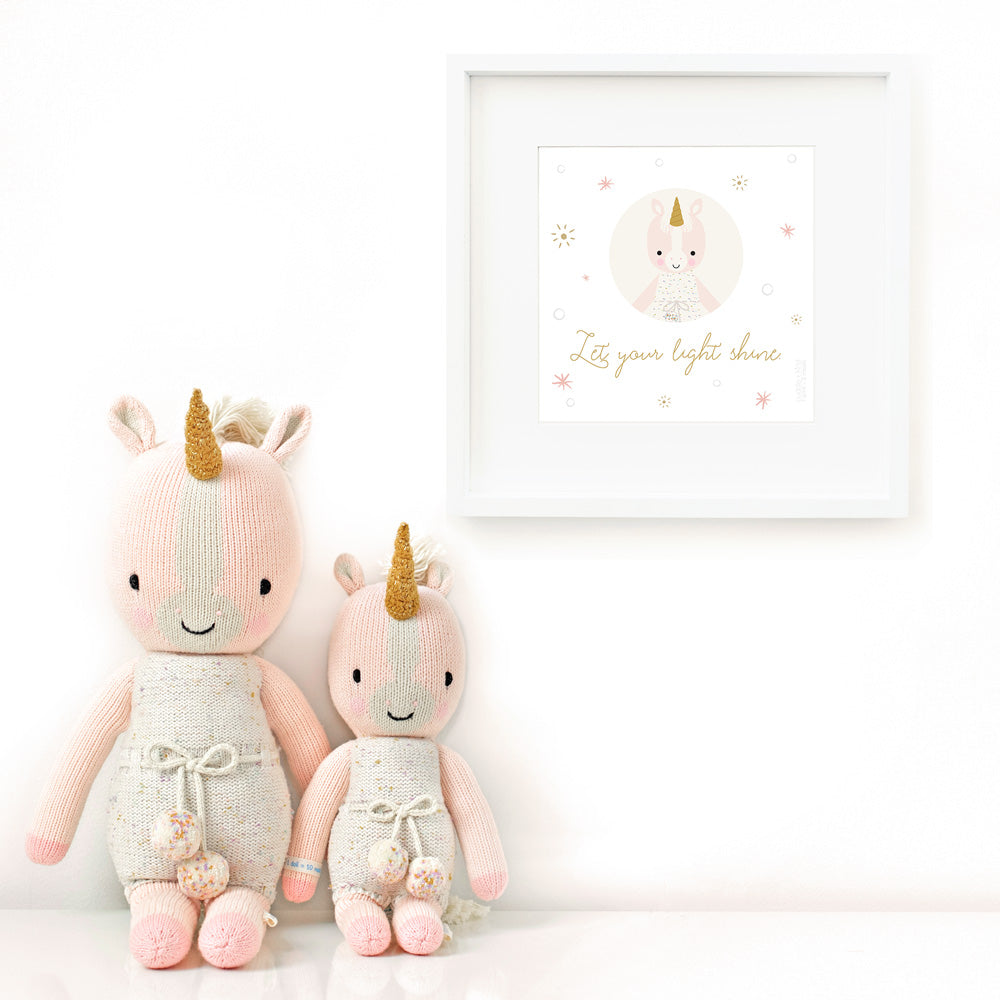 Two Ella the unicorn stuffed dolls sitting beside a framed print with a picture of Ella that says “Let your light shine.”