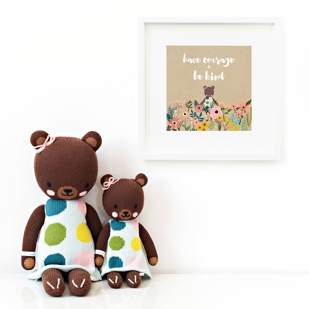 Two Ivy the bear stuffed dolls sitting beside a framed print with a picture of Ivy that says “Have courage + be kind.”