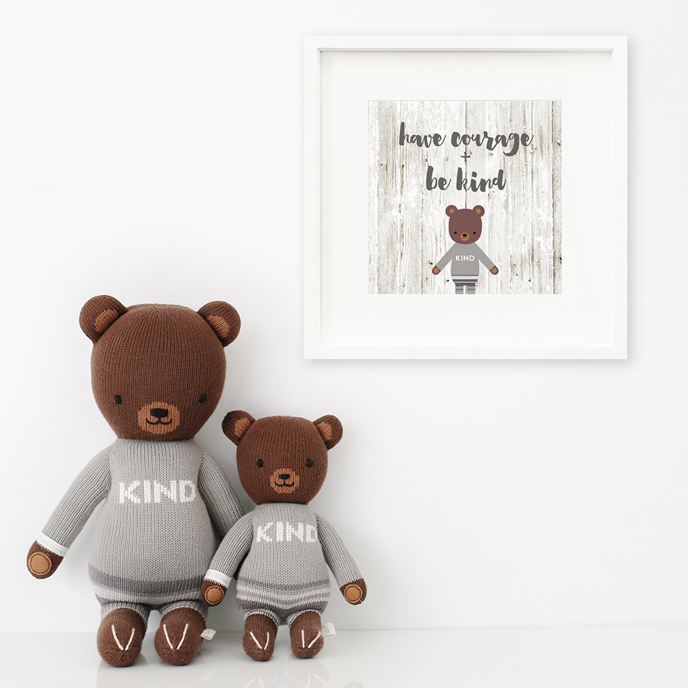 Two Oliver the bear stuffed dolls sitting beside a framed print with a picture of Oliver that says “Have courage + be kind.”
