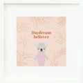 An inspirational print with a graphic of Claire the koala on a blush-colored background decorated with leaves and the words “Daydream believer” in orange-pink.