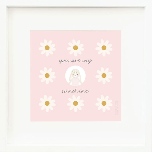 An inspirational print with a graphic of Hannah the bunny in blush, on a pink background decorated with daisies and the words “You are my sunshine” in dark gray.